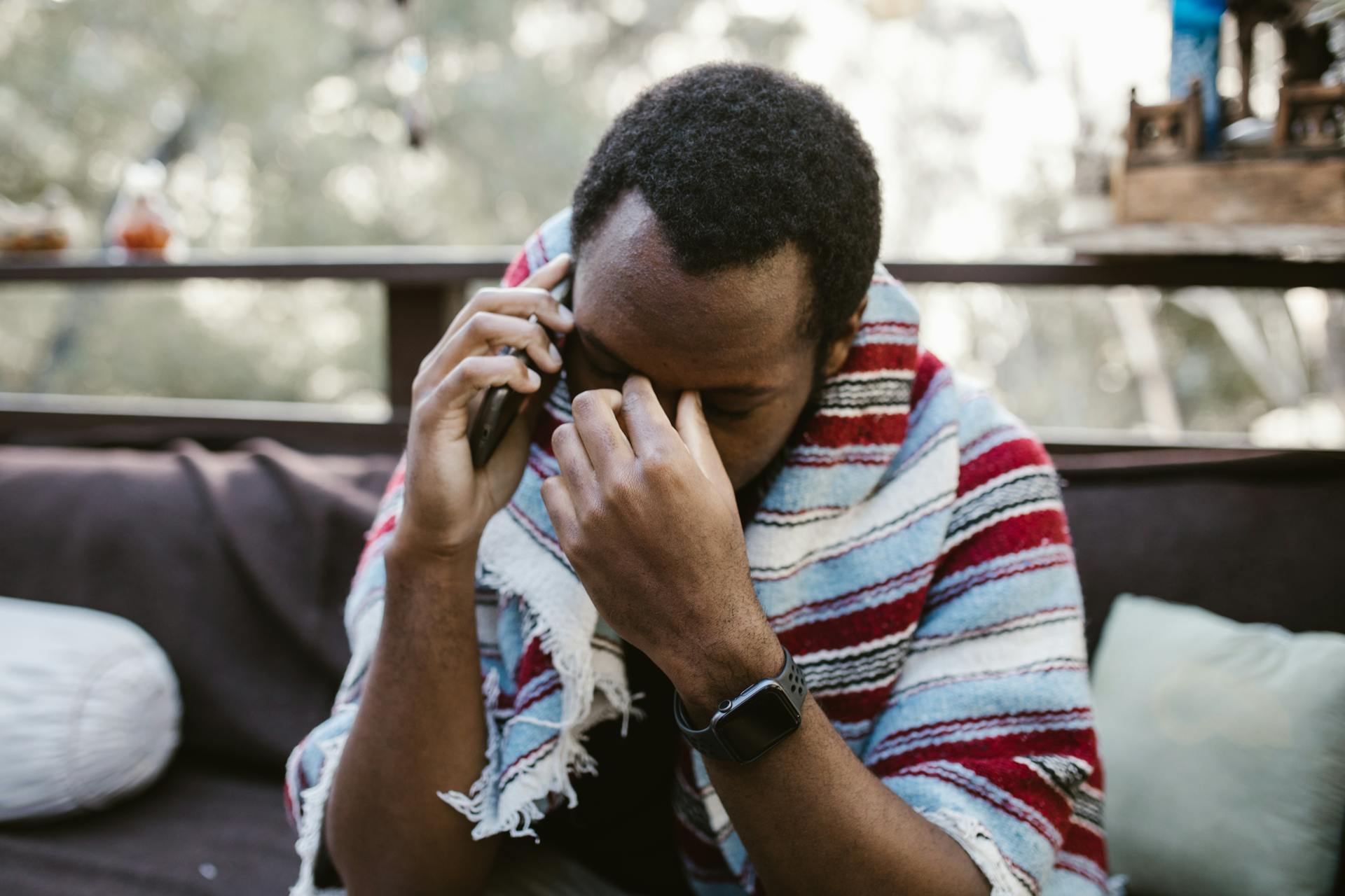 A Black man with a blanket on his shoulders sits on a couch and makes a phone call