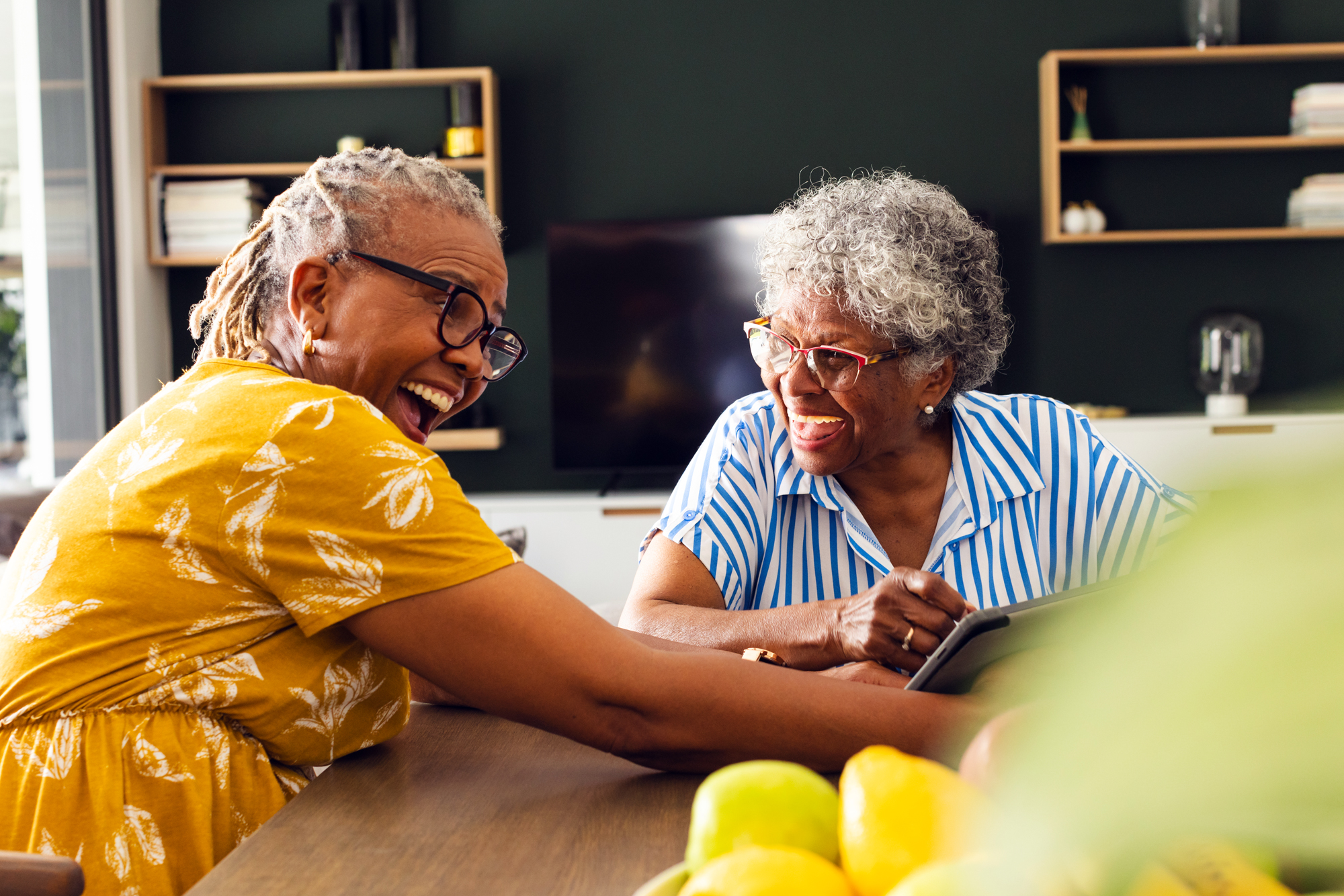 Two black women who are seniors sit at a table and laugh while looking at a tablet.